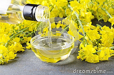 Pouring canola oil into the glass bowl against rapeseed blossoms on the grey surface, closeup shot Stock Photo