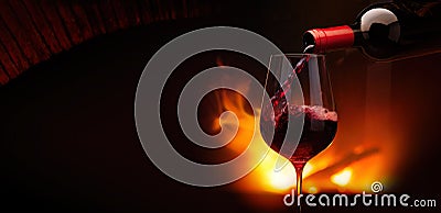 Pouring red wine into a glass at night near fireplace flames. Cozy wintertime background with copy space Stock Photo