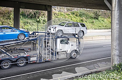 Powerful big rig car hauler semi truck transporting cars on semi trailer running under the bridge over the wide highway road Stock Photo