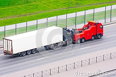 Powerful big rig semi truck tractor tows with attached broken evacuated truck with container trailer driving on city highway, Stock Photo