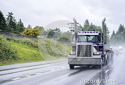 Powerful classic big rig blue semi truck with flat bed semi trailer running on the wet raining road in front of another traffic Stock Photo