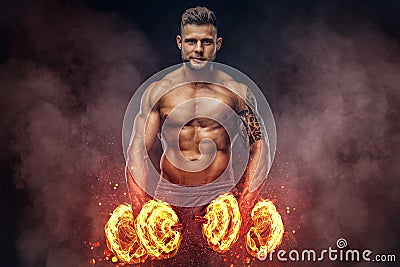 Handsome shirtless tattooed bodybuilder with stylish haircut and beard, wearing sports shorts, posing in a studio. Fire Stock Photo