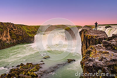 Powerful waterfall Godafoss at beautiful red sunset with a lonely traveler standing at its cliff, Iceland, summer, scenic view Stock Photo