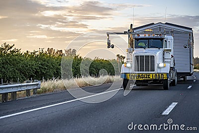 Powerful white big rig classic semi truck tractor with oversize load sign on the bumper transporting oversized manufactured house Stock Photo