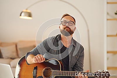 Practicing songwriting skill. Man in casual clothes and with acoustic guitar is indoors Stock Photo