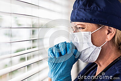 Prayerful Stressed Female Doctor or Nurse Crying At Window Wearing Medical Face Mask Stock Photo