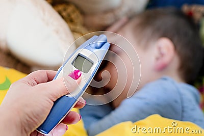 Pre-school sick boy lying on pillow in bed. Mother is measuring body temperature on his forehead with modern digital thermometer Stock Photo