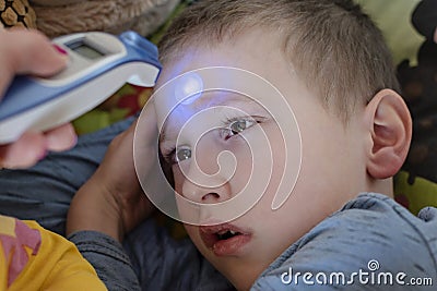 Pre-school sick boy lying on pillow in bed. Mother is measuring body temperature on his forehead with modern digital thermometer Stock Photo