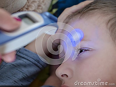 Pre-school sick boy lying on pillow in bed. Mother is measuring body temperature on his forehead with modern digital thermometer Stock Photo