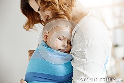 Precious little newborn boy having deep sleep at day on mother chest in blue baby sling. Mom kissing baby head and Stock Photo