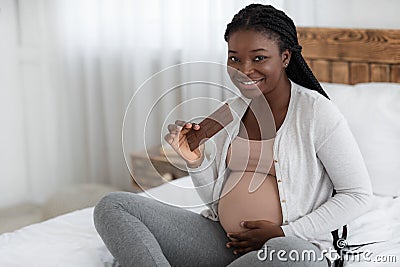 Pregnancy Cravings. Smiling Expectant Black Woman Eating Milk Chocolate Bar At Home Stock Photo