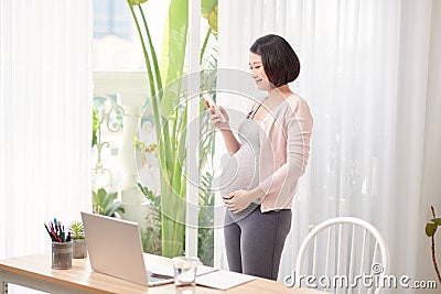 Pregnant asian woman working from home in modern office contacting using mobile phone in call talking Stock Photo