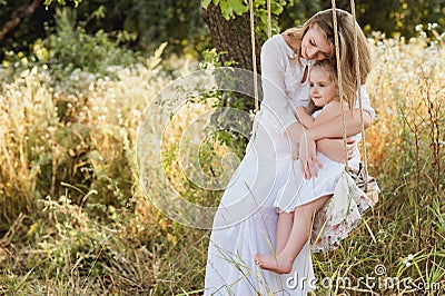 Pregnant beautiful mother with little blonde girl in a white dress sitting on a swing, laughing, childhood, relaxation Stock Photo