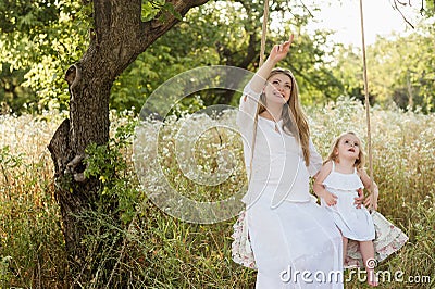 Pregnant beautiful mother with little blonde girl in a white dress sitting on a swing, laughing, childhood, relaxation, serenity, Stock Photo