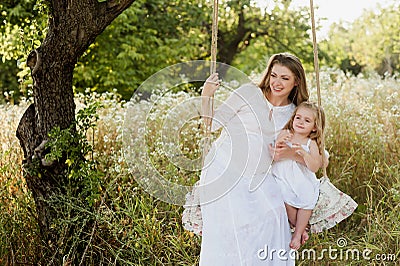 Pregnant beautiful mother with little blonde girl in a white dress sitting on a swing, laughing, childhood, relaxation, serenity, Stock Photo