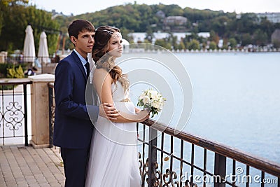 A pregnant bride in a wedding dress is standing on the dock, admiring the view of the river, behind her hugs her husband Stock Photo