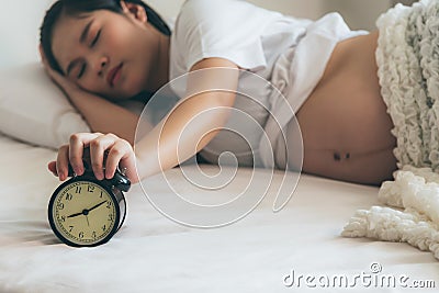 Pregnant teen woman lying in a white bed, is using her hands Press stop noise from alarm clock Stock Photo