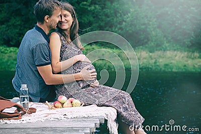 Pregnant woman and her husband sitting together on wooden pier on the lake. Candid photo of expecting baby in young family. Happy Stock Photo