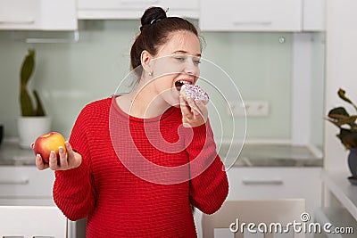 Pregnant woman in kitchen holding apple in hand and eating cake. Young attractive female decides to eat unhealthy food, looks Stock Photo