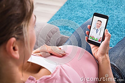 Pregnant Woman Sitting On Sofa Video Conferencing With Doctor Stock Photo