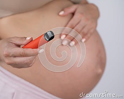 A pregnant woman smokes a vape. A girl holds an electronic cigarette against the background of her bare tummy. Stock Photo