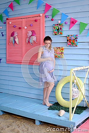 Pregnant young woman standing in blue dress near children houe and holding belly. Stock Photo