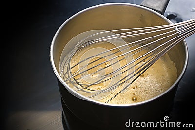 Preparation of hollandaise sauce, egg mixture is whisked at low temperature until foamy in a steel pot on a black cooktop, cooking Stock Photo