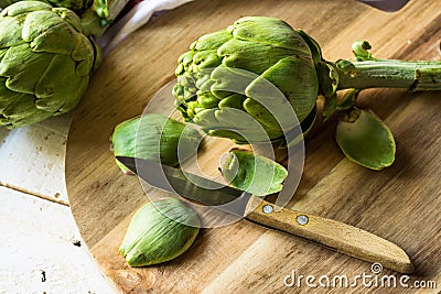 Preparing and peeling fresh artichoke for cooking, scattered green petals, cutting board, knife Stock Photo