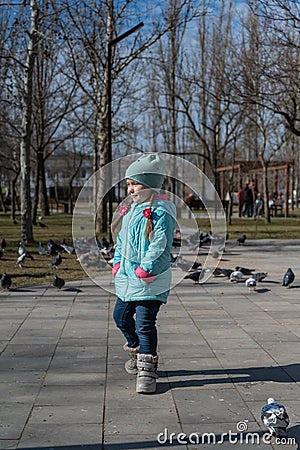 Preschool-aged girl in a blue jacket twirls around herself in front of a flock of city doves. Portrait of a five-year-old child Stock Photo