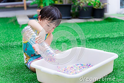 Preschool child boy is using two hands to hold plastic bottle to pour water beads or rainbow beads into white basin. Stock Photo