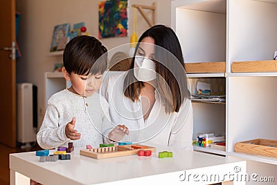 Preschooler Kid playing tetris wood puzzle with teacher educador help using face mask for coronavirus pandemic. Homeshooling. Stock Photo