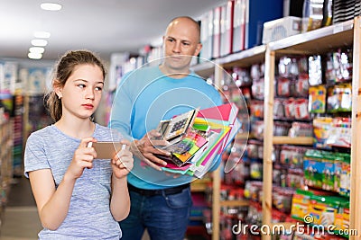 Preteen girl absorbed in phone while father showing school stationery to her in store Stock Photo