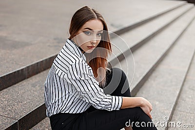 Pretty American young woman in fashionable jeans in a vintage black and white striped blouse with elegant velvet necklaces sits Stock Photo