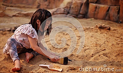 A young Asian girl playing in a sandbox with a modeled dinosaur fossil using brush and shovel Stock Photo