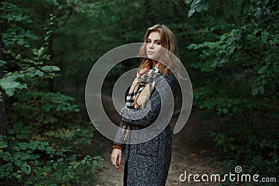 Pretty attractive european young woman in a fashionable checkered scarf in a chic gray coat walks in the woods near green shrubs. Stock Photo