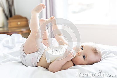 Pretty baby girl drinks water from bottle lying on bed. Child weared diaper in nursery room. Stock Photo