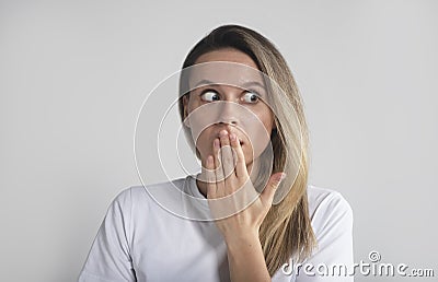 Pretty blond girl in shock and surprise closing mouth with hand looking aside over grey studio background Stock Photo