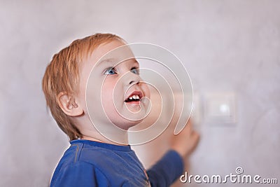 Pretty blonde caucasian baby boy turns on/off the light-switch, looking up. Big blue eyes, close up portrait, copy space, indoors. Stock Photo