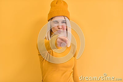 Pretty blonde female gives thumb up, approves new good idea, blink with the eye, wears yellow outfit, stands against yellow studio Stock Photo