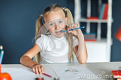 Pretty blondy girl thinking while doing her homework and holding a blue pencil, at home table. Education and school concept Stock Photo