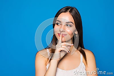 Pretty charming young woman having secret while holding finger on lips and showing silence sign while standing on blue background Stock Photo