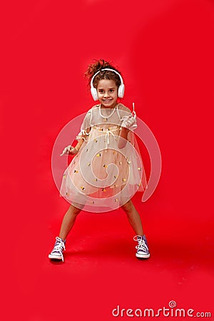 Pretty cool ittle curly girl having fun and listens to music in the white headphones and dancing over red background Stock Photo