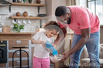 Pretty curly-haired kid pouring flour and looking happy, her father helping Stock Photo