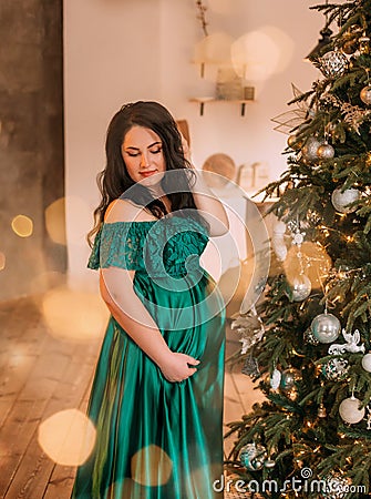 Pretty dark-haired pregnant woman in gorgeous adorable green emerald long dress near New Year tree, lady modestly Stock Photo