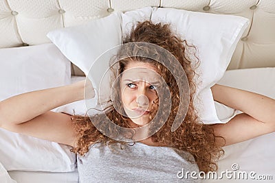 Pretty discontent female with curly covers ears with pillow, lies in bed, has insomnia, cant fall asleep because of noise, has sle Stock Photo