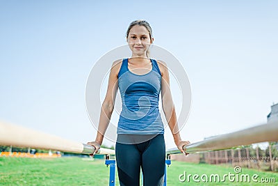 A pretty girl in a blue shirt and leggings doing sports on uneven bars looking at camera and smiling. Stock Photo