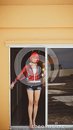 Pretty girl in devil costume standing in a gloomy corridor by the window. Wears a red wig with horns, denim shorts and Stock Photo