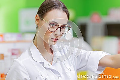 A pretty graceful dark-haired lady with glasses, dressed in a white coat, carefully examies the goods on the shelf in Stock Photo