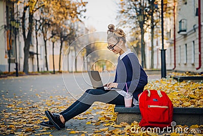 Pretty hipster teen girl sitting on a sidewalk on autumn city street and working laptop computer. Schoolgirl using Stock Photo