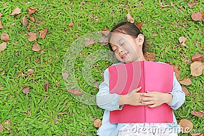 Pretty little girl with book lying on green grass with dried leaves in the summer garden Stock Photo